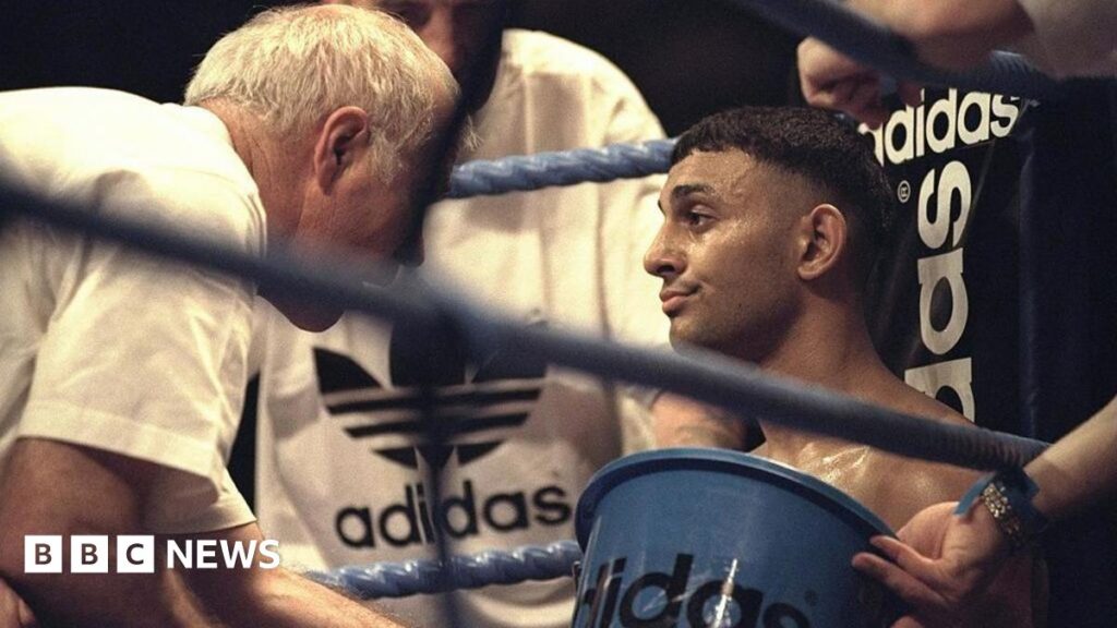 A boxer sits in the corner of a boxing ring, being attended to by his team - one holding an Adidas-branded bucket and others dressed in Adidas gear - offering support and guidance between rounds.