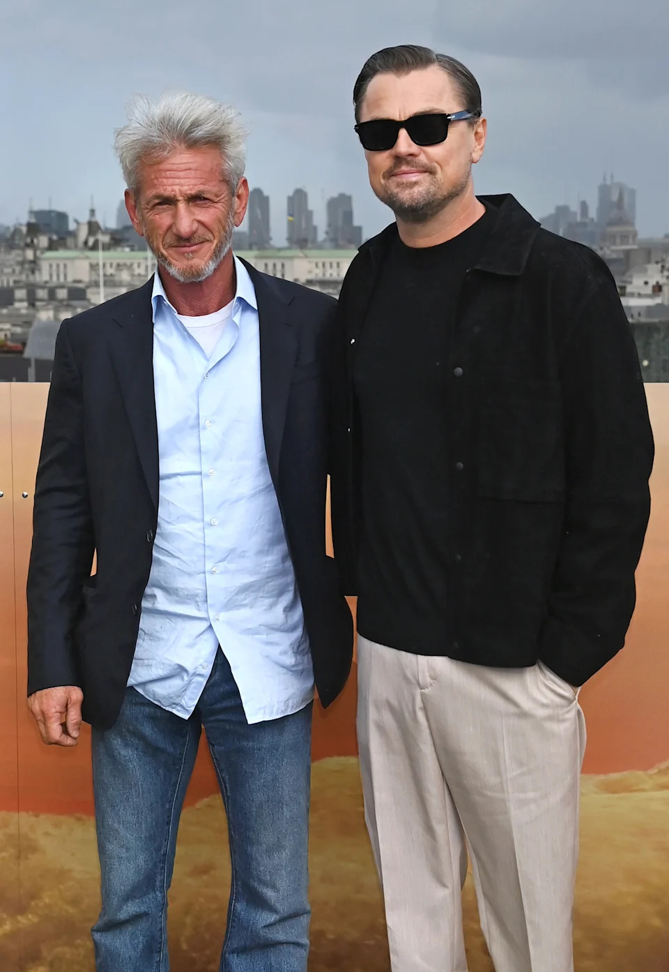 Sean Penn and Leonardo DiCaprio pose together, with city buildings in the background.