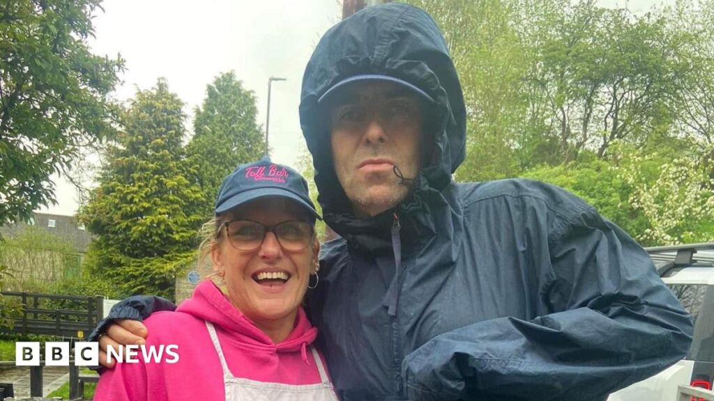 Liam Gallagher in a waterproof coat with arm round woman in lilac top