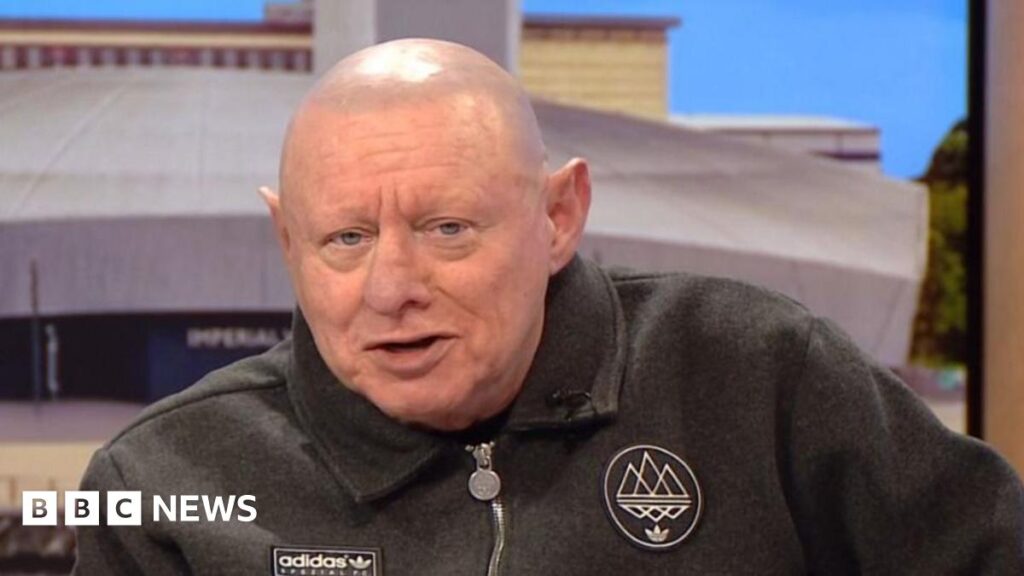 Shaun Ryder missed Mani's funeral as pneumonia led to collapse – BBC Shaun Ryder speaking on BBC Breakfast. He wears a grey adidas zipped jumper and is bald with blue eyes.