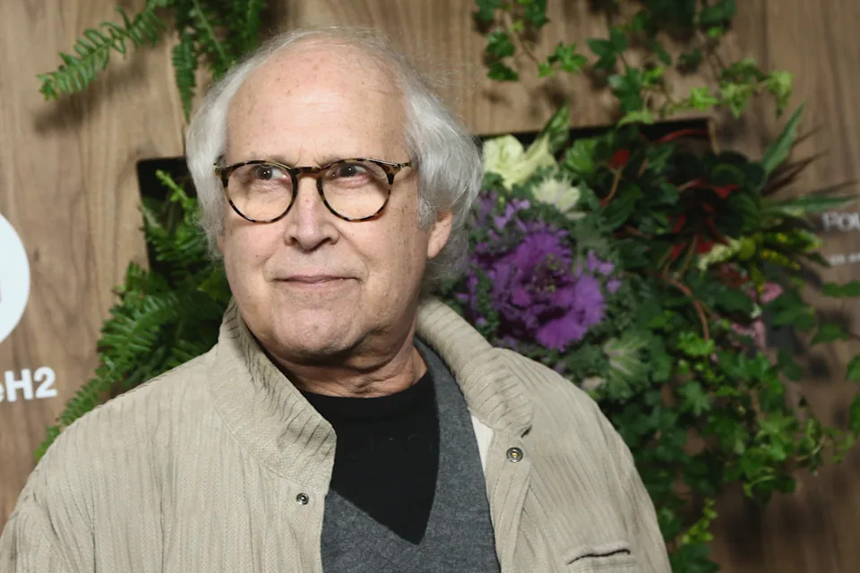 A person wearing glasses and a casual coat with a sweater underneath, standing in front of a floral backdrop