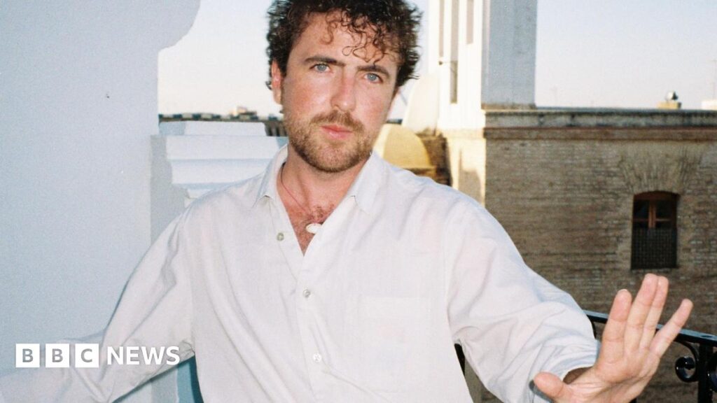 Michael McGovern - a man with dark curly hair, a beard, goatee and a white shirt, standing outside. He is holding one hand up ahead of himself.