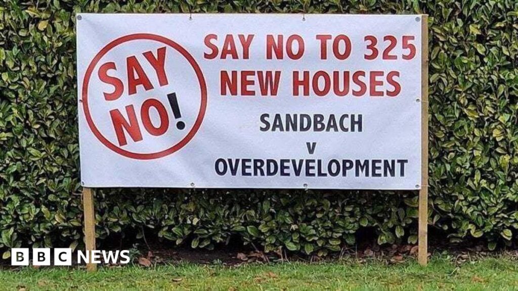 A large white banner alongside a hedge with red and black lettering which reads "Say no to 325 new houses - Sandbach v overdevelopment"