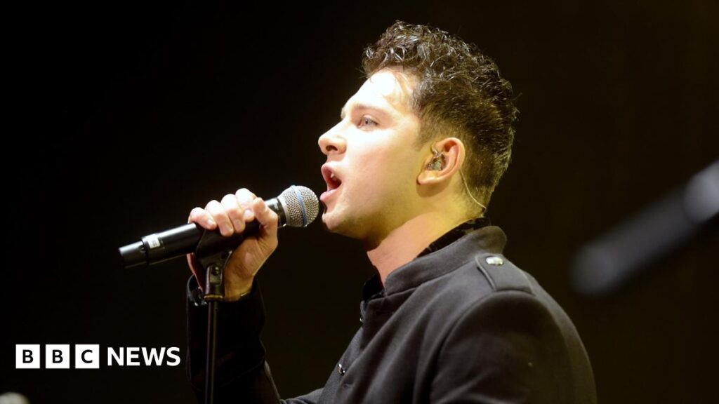 Man with brown hair sings into microphone on stage with red lighting shining down on him. He wears a tan t-shirt and black punk wristband.