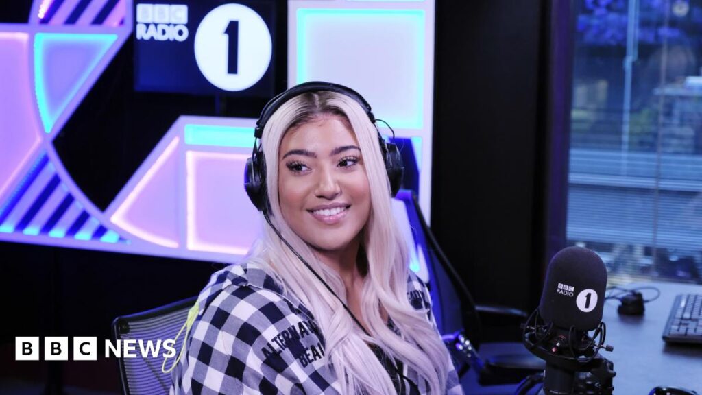 Radio 1Xtra’s first female Rap Show host leaves the station Tiffany Calver is sitting in a studio in front of a BBC Radio 1 microphone. She has headphones on is wearing a black-and-white checked jacket and has blonde hair.