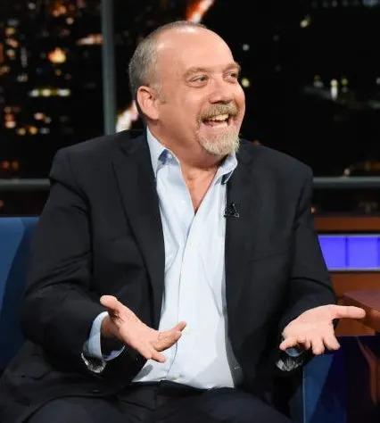 Two men in suits are laughing and talking on a late-night talk show set, with a night cityscape visible in the background