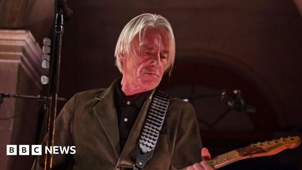 Paul Weller completes Ludlow Castle summer concerts line-up A man with long grey hair, a brown jacket and a brown guitar performs on stage. He looks towards the head of his guitar while a black microphone stand can be seen in front of him.