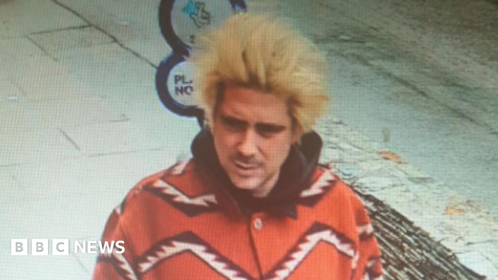 Alexander Key is walking in a street. He has long peroxided hair standing up and a moustache. He is wearing a bright red coat which has a bold zigzag print in white and black.