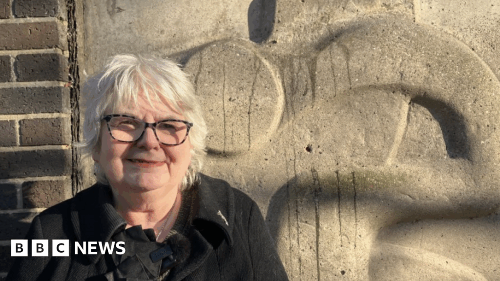 Renewed call to save Grimsby car park art as demolition begins Angela Greenfield - a woman with short, grey hair, smiling. She is wearing dark glasses and a black coat. She is standing in front of a stone-coloured sculpture which is on the side of a car park