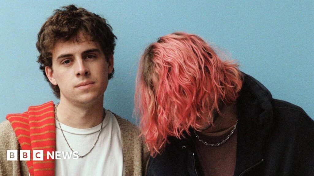 Press photo of Royel Otis standing against a plain blue background. Guitarist Royel Maddell covers his face with long, pink tousled hair