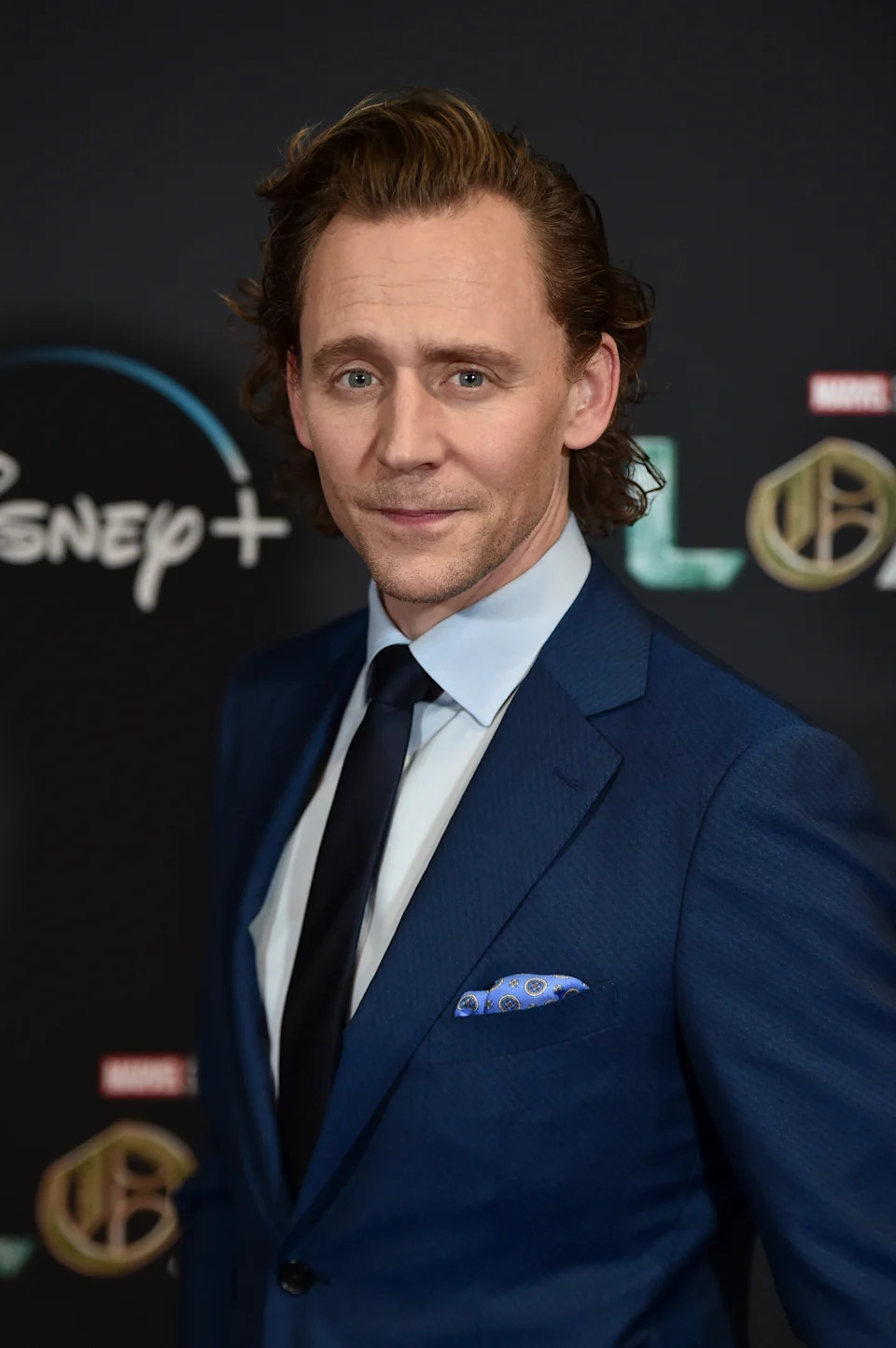 Actor in a formal dark suit and tie poses at a Disney+ event backdrop, conveying elegance and sophistication