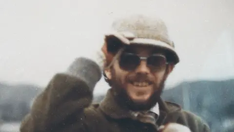 Cwmni Da/S4C Kenner Elias Jones is a white man in his 30s or 40s in this picture, with a brown beard and moustache. He wears dark lensed prescription glasses and a cream coloured hat which he is holding, a checked shirt and tie covered by a brown coat with a collar. He is smiling at the camera. The image is grainy.