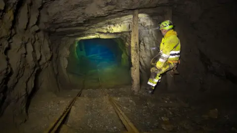 Tim Clarke A man is a yellow boilersuit stands in an old mine working, the rocky tunnel ends in a green blue pool of water 