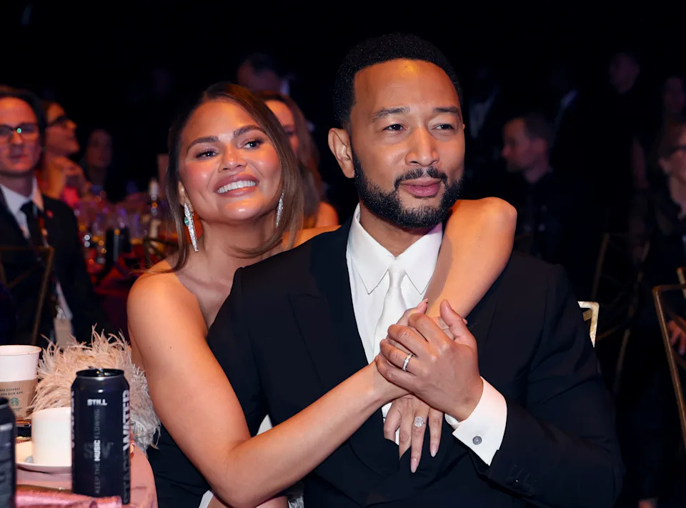 Seated at a dinner table, Teigen drapes her arms over her husband at the MusiCares gala.
