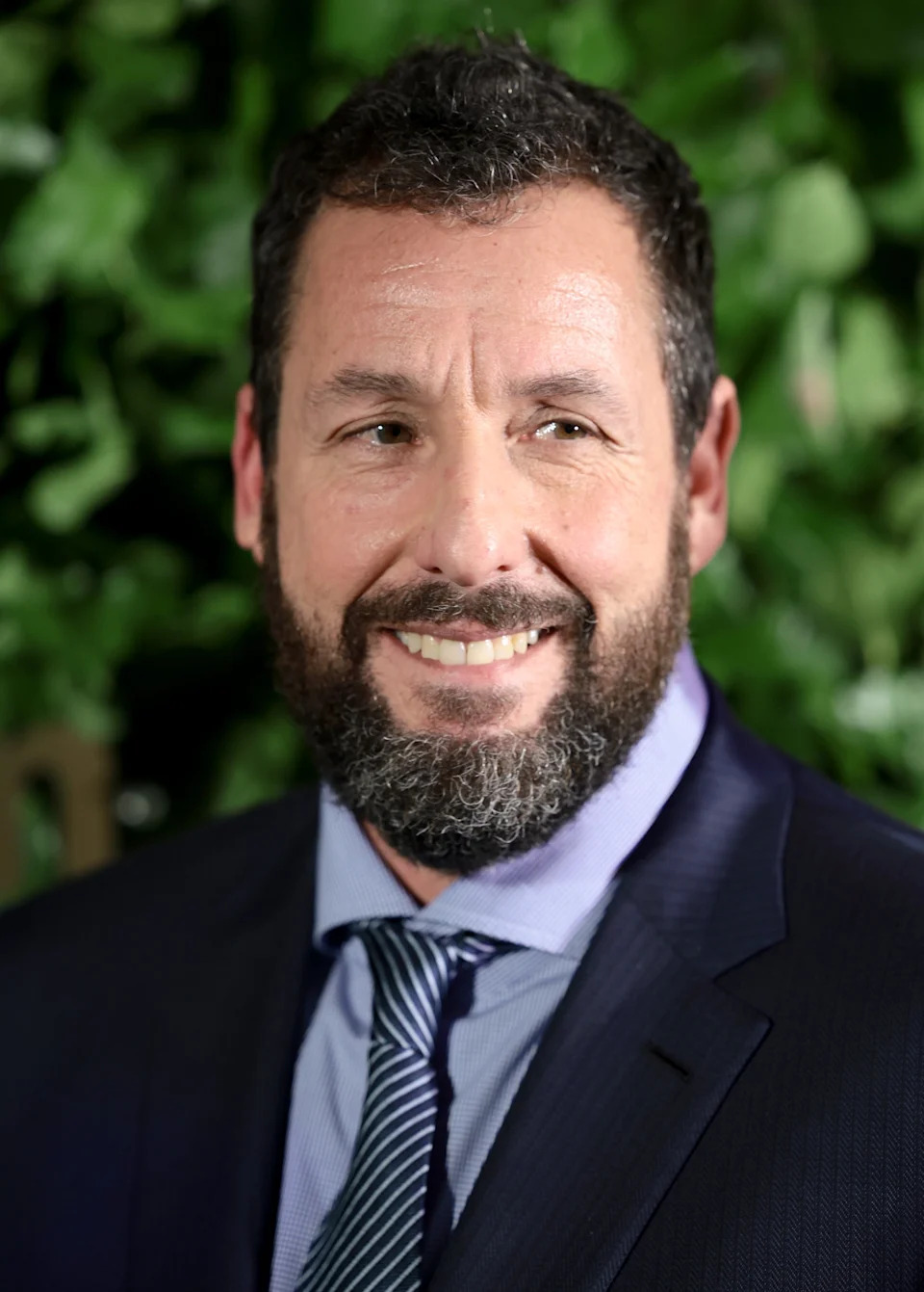 A person with a beard, wearing a suit and tie, smiles in front of a leafy backdrop at an event