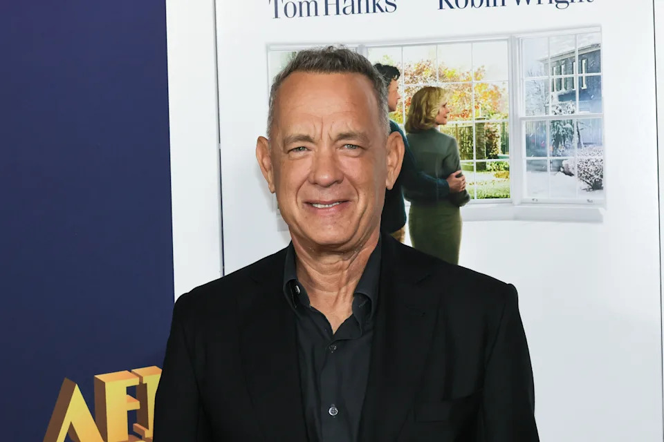 A person in a dark suit smiles at a film premiere, standing in front of a movie poster with two people looking out a window