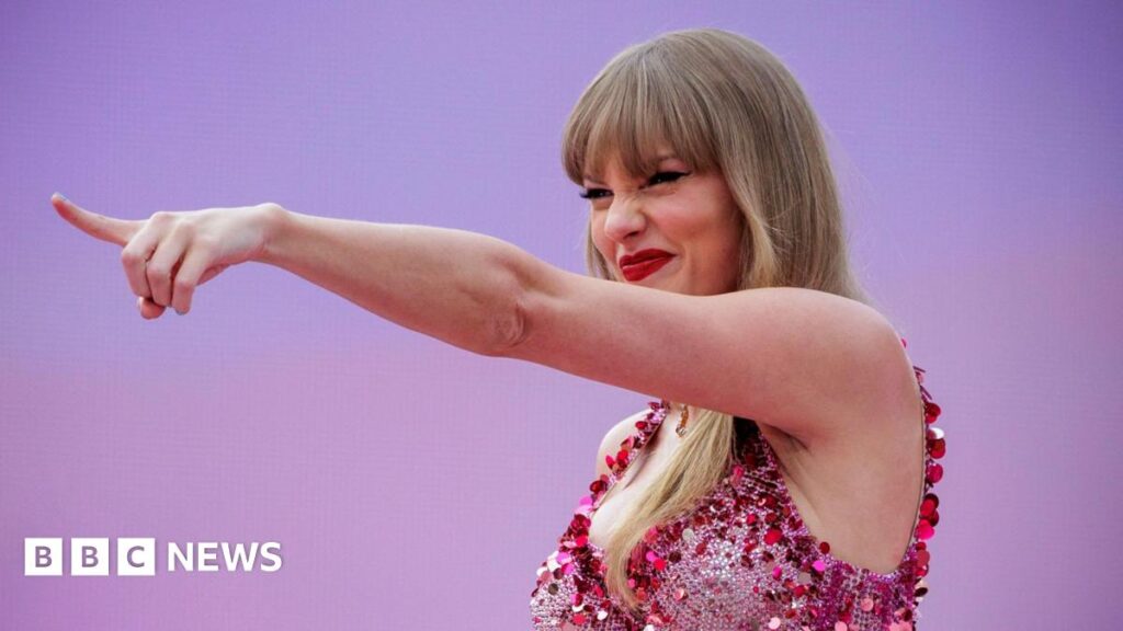 Taylor Swift inducted into Songwriters Hall of Fame Taylor Swift points into the distance with a grin and her eyes squinted beneath her blonde fringe. She wears a sparkly pink fitted bodice and red lipstick. The background is pinkish-purple gradient and matches her outfit.