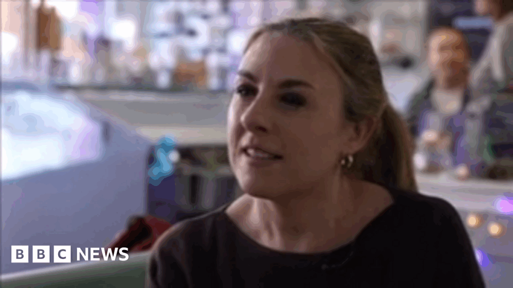 Alex Hoskyn has light brown hair in a ponytail. She is wearing a dark jumper and is sitting in a cafe