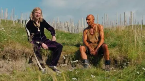 Sony Pictures Jack O'Connell, dressed as his character Sir Jimmy Crystal, speaks with Ralph Fiennes' character Dr Kelson in a scene in the new film, 28 Years Later: The Bone Temple. O'Connell is wearing a dirty tracksuit and chains around his neck, and has a blonde mullet. Bald Fiennes is wearing a raggedy vest, shorts and boots.