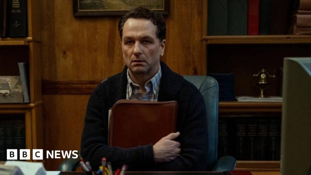 Apple TV’s Widow’s Bay star Matthew Rhys plays mayor of cursed town Actor Matthew Rhys sits in a chair at a wooden desk holding a folder to his chest. On the table is a black landline telephone, a rolodex, a computer and a pot of pens. He stares into the distance with a grave look on his face.