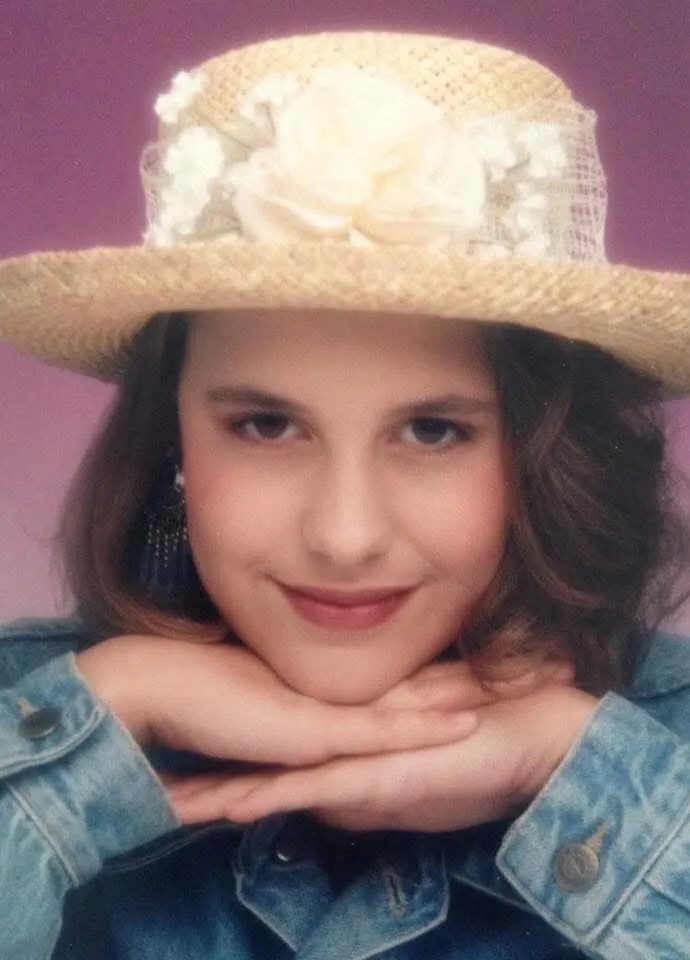 A young girl poses for the camera with her hands under her chin. She is wearing a jean jacket and a straw hat.