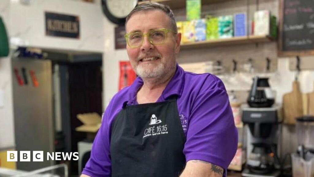 Rushden cafe for homeless charity ready to expand operations Stan Robertson, standing in a cafe, wearing a purple shirt, with a black apron, with cafe equipment behind him. He is smiling, with yellow-framed glasses on, a grey beard and short greying hair.