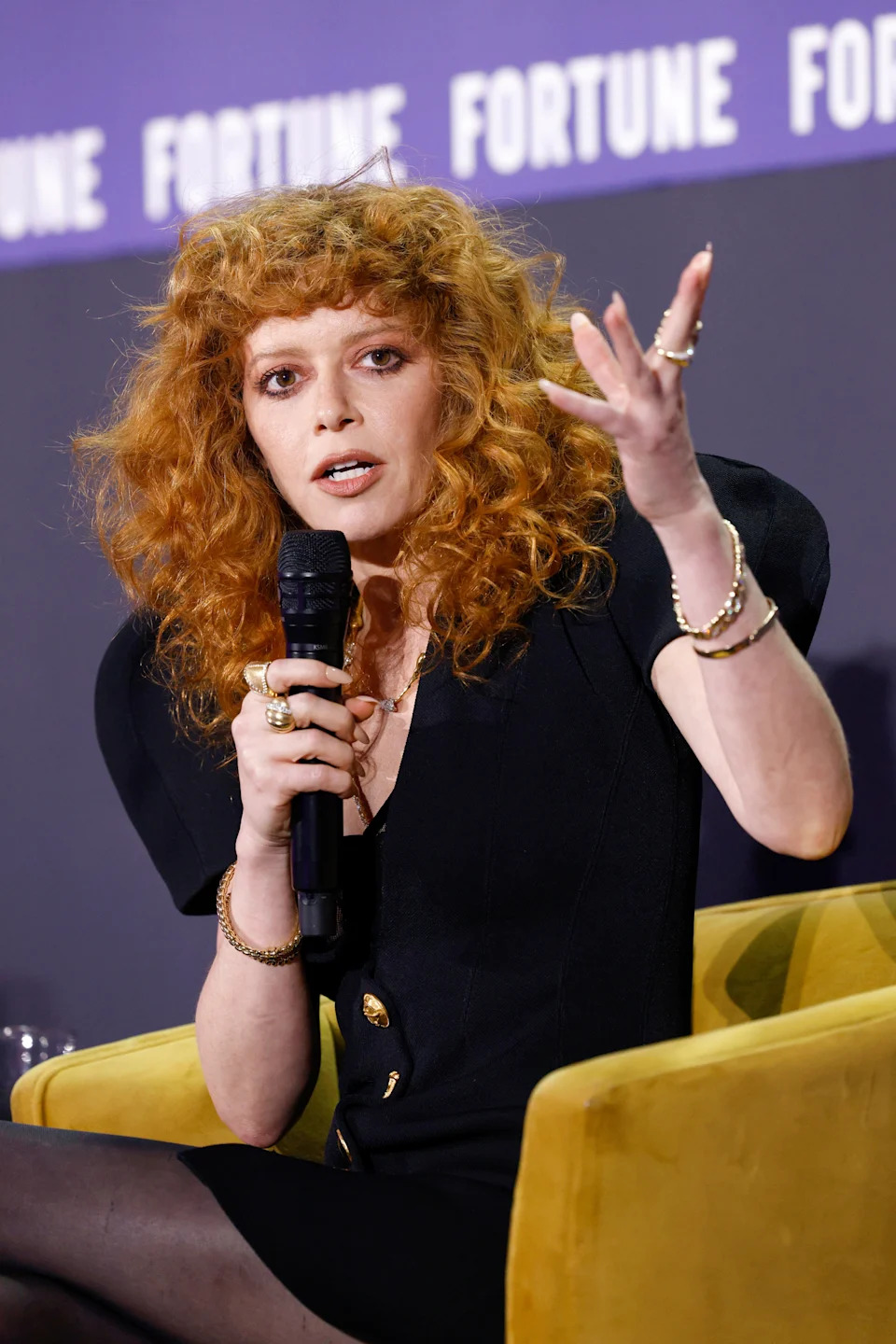 Natasha Lyonne with curly hair speaks into a microphone on a panel, wearing a stylish outfit with padded shoulders and gold accessories