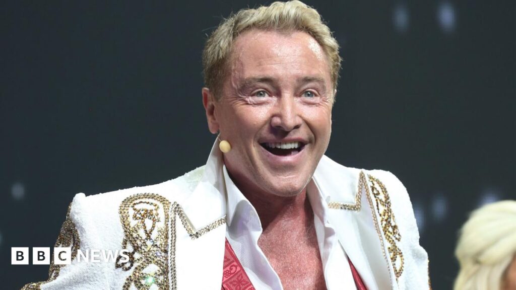 Michael Flatley is wearing a black headband, a white open jacket with black detailing and black trousers with white detailing. Behind him are some dancers in darkness.
