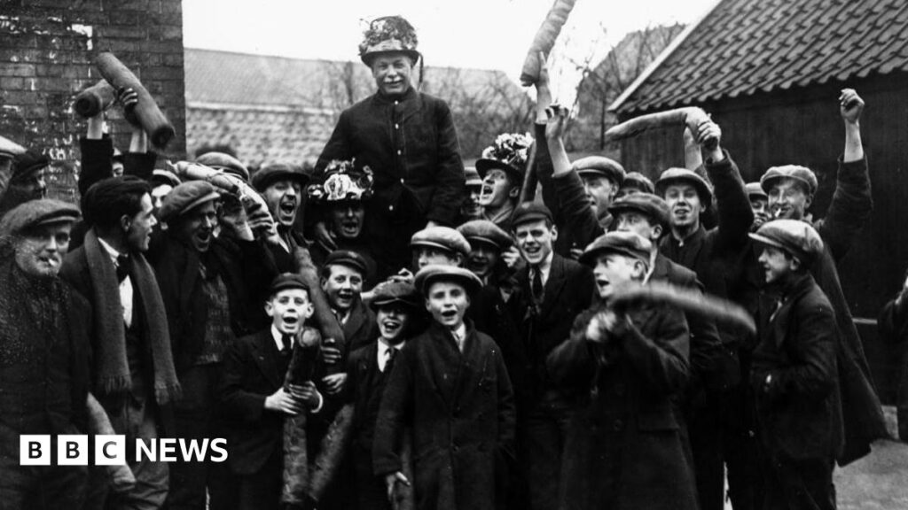 Children help keep centuries old Haxey Hood tradition alive A black and white photo of several young boys in caps and suits waving rolls of leather about three foot long above them. In the middle you can see an old man wearing a garlanded top hat being held up by three other men who are shouting with their mouths wide open, also wearing garlanded top hats. Brick buildings can be seen behind them.