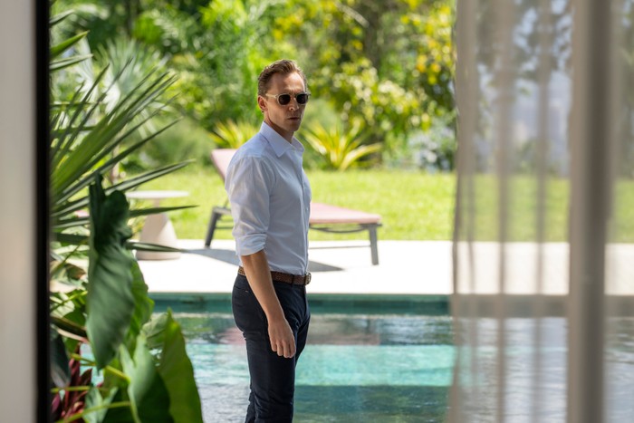The Night Manager S2 Tom Hiddleston in The Night Manager season 2, stood by an outdoor pool, wearing a shirt, trousers and sunglasses.