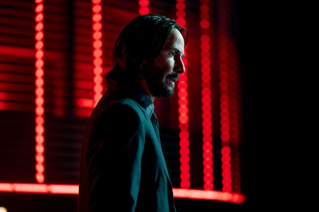 Keanu Reeves as John Wick in the movie John Wick: Chapter 4, in a dark space with red neon lights behind him. He is wearing a dark suit.