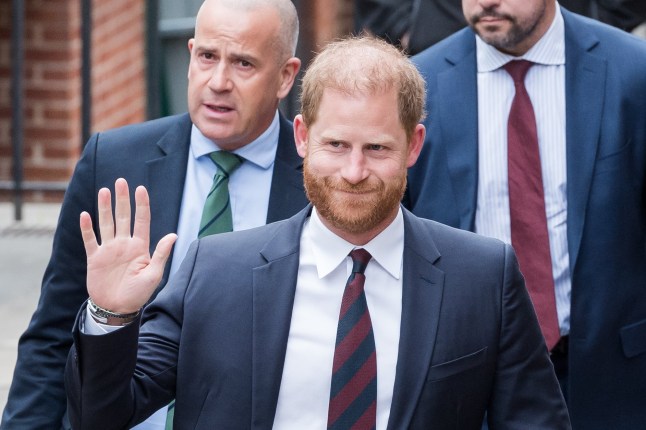 Prince Harry dressed in a suit and waving as two men walk behind him