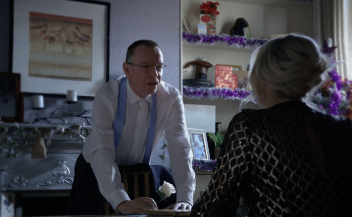 Adam Woodyatt as Ian Beale in a wedding suit leaning on a kitchen table looking at Gillian Taylforth as Kathy Beale in EastEnders.