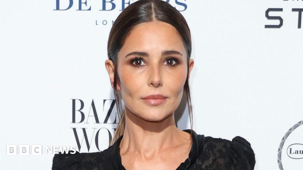 Cheryl posing for photos at an event against a white backdrop with brand names printed on