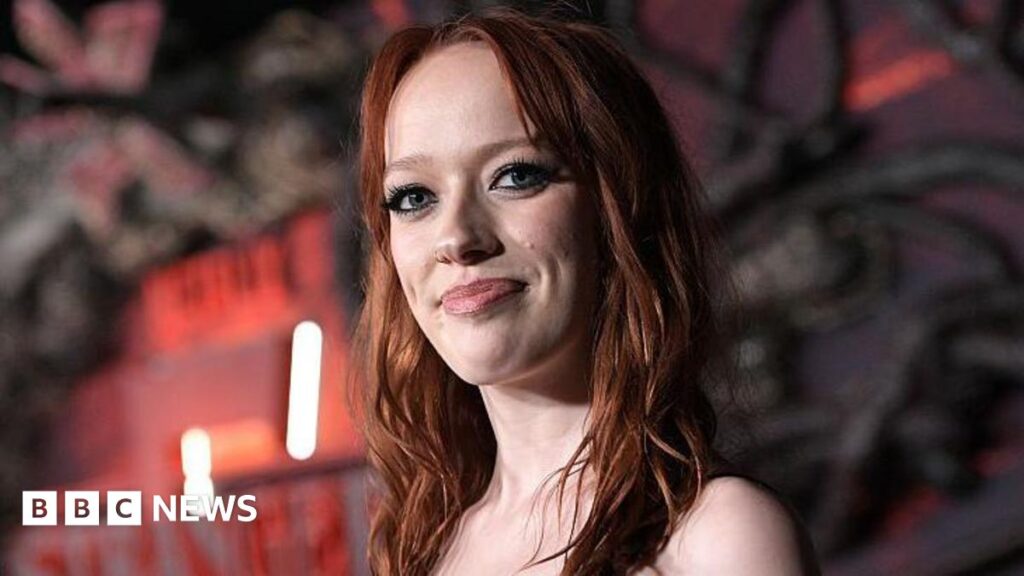 Marybeth is pictured at a film premier. There is a black and red background with strobe lighting. She is smiling at the camera and has ginger hair.