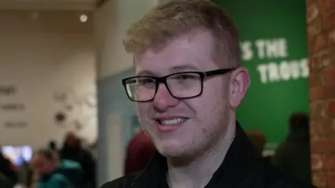Morgan Clossick, a young man wearing black-framed glasses, smiles while standing by part of a museum exhibit. People can be seen milling around behind him. 