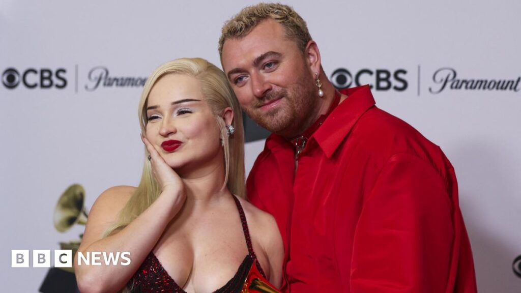 A woman with blonde hair and red lipstick poses with her hand cupping her chin on a red carpet. Standing next to her is Sam Smith who has short hair and a pearl dangling earring. They are wearing a red shirt and has one hand round her waist, with their other holding a small gold trophy.