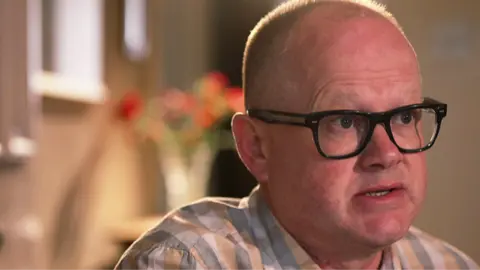Cwmni Da/S4C Marc Edwards is a white man with balding, fair closely cropped hair. His head and shoulders are visible. He wears black, heavy-rimmed glasses and a grey, white and tan checked shirt. He is looking towards the right of the image and appears to be talking seriously. The background is blurred but it is a room and a vase of flowers on a shelf can be discerned. 