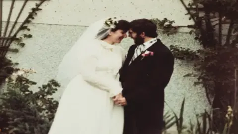 Cwmni Da/S4C Lee McKenzie is a white woman with dark hair and is wearing a white long-sleeved wedding dress and a veil. Kenner Jones is wearing a black morning suit with a black, grey and white striped tie and a pink flower on the lapel. They are standing in front of a white wall with plants to either side. They are holding hands and smiling at each other.