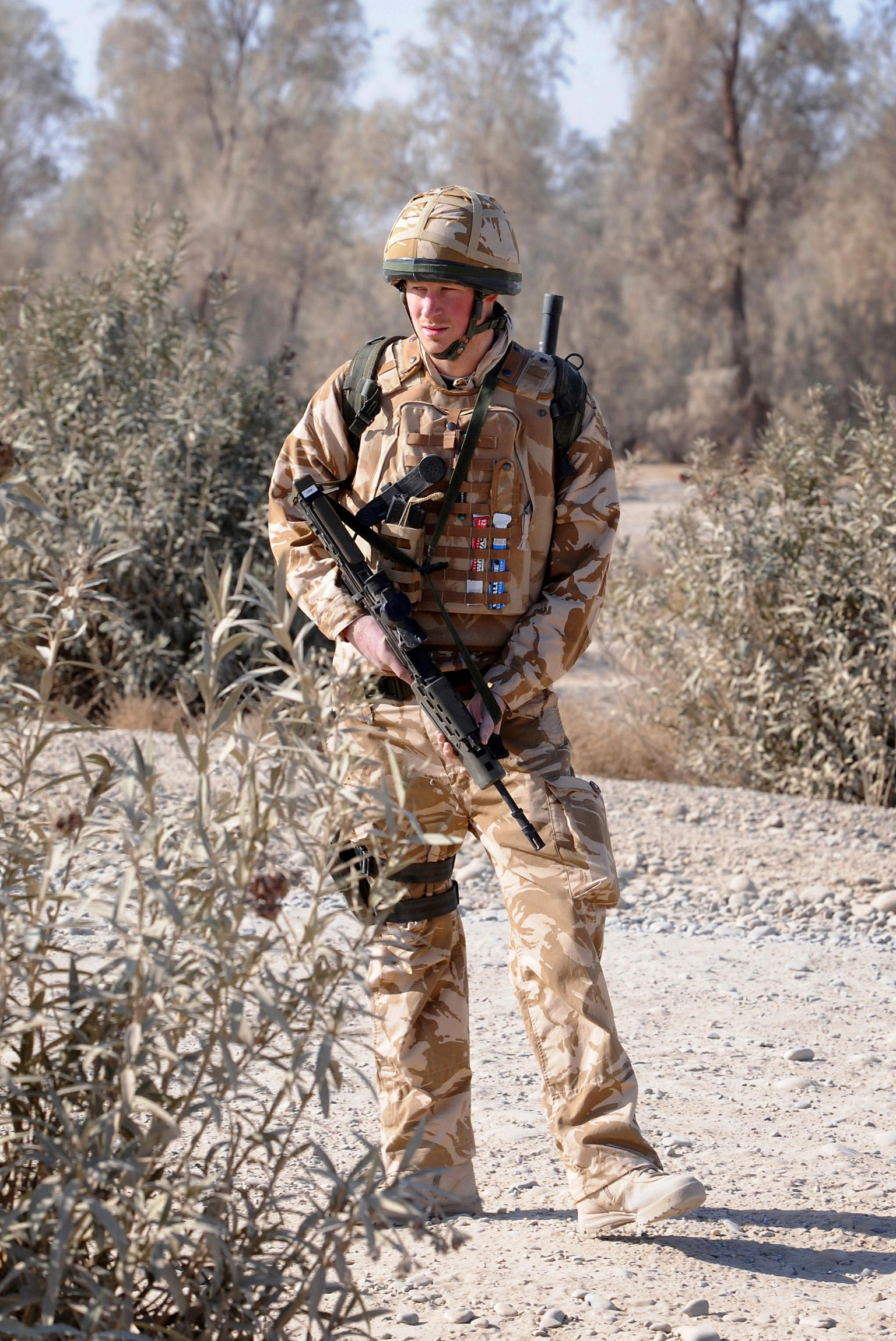 Prince Harry wearing a military uniform walking in Afghanistan with a gun
