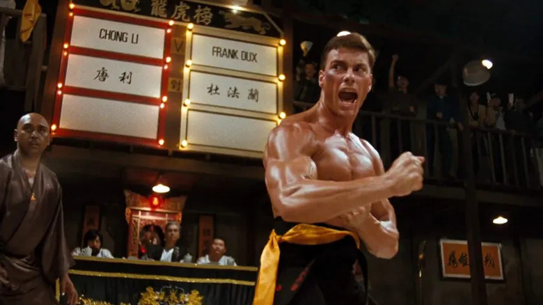 Frank Dux (Jean-Claude Van Damme) shouts while striking a pose in front of a scoreboard in Bloodsport
