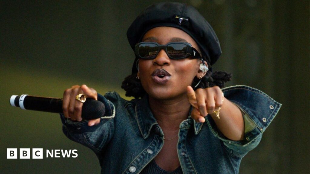 Little Simz performs on stage wearing a denim jacket, black beret and pitch black sunglasses. She is pointing to the audience and encouraging them to sing along.