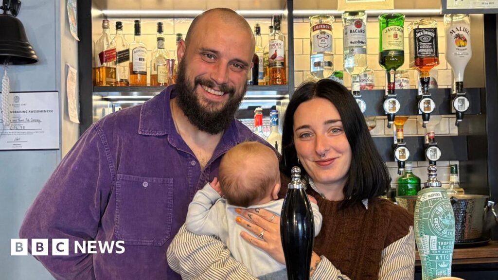 Three people including a baby standing at the bar of a pub. Behind them are rows of bottles of alcohol and some on optics.