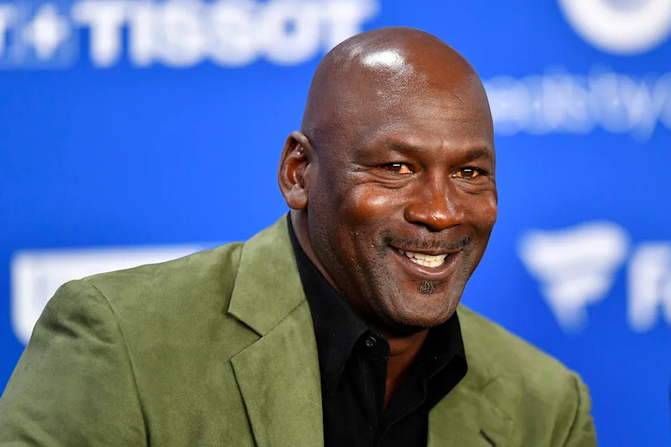 A man smiles at an event, wearing a green blazer over a black shirt with a blue backdrop displaying sponsor logos