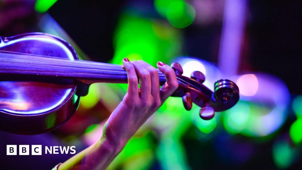 A close up image showing part of a female musicians hand and arm as she holds a fiddle. There are blurry bright lights in the background.