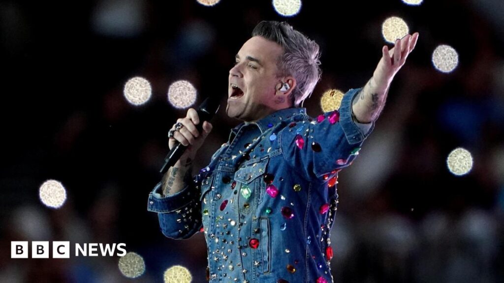 Robbie Williams singing into a microphone. He is wearing a jean shirt with a white collar and has grey spiky hair.