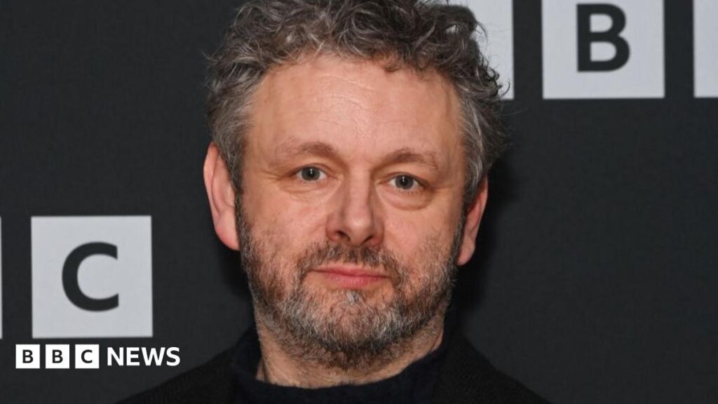 Michael Sheen fronts investigation series into chemical contamination in Wales Michael Sheen has a grey beard and long hair. He wears a blue knitted jumper and a green jacket