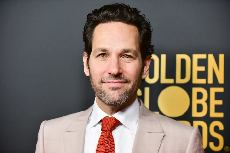 A man in a suit with a white shirt and red tie stands in front of a "Golden Globe Awards" backdrop