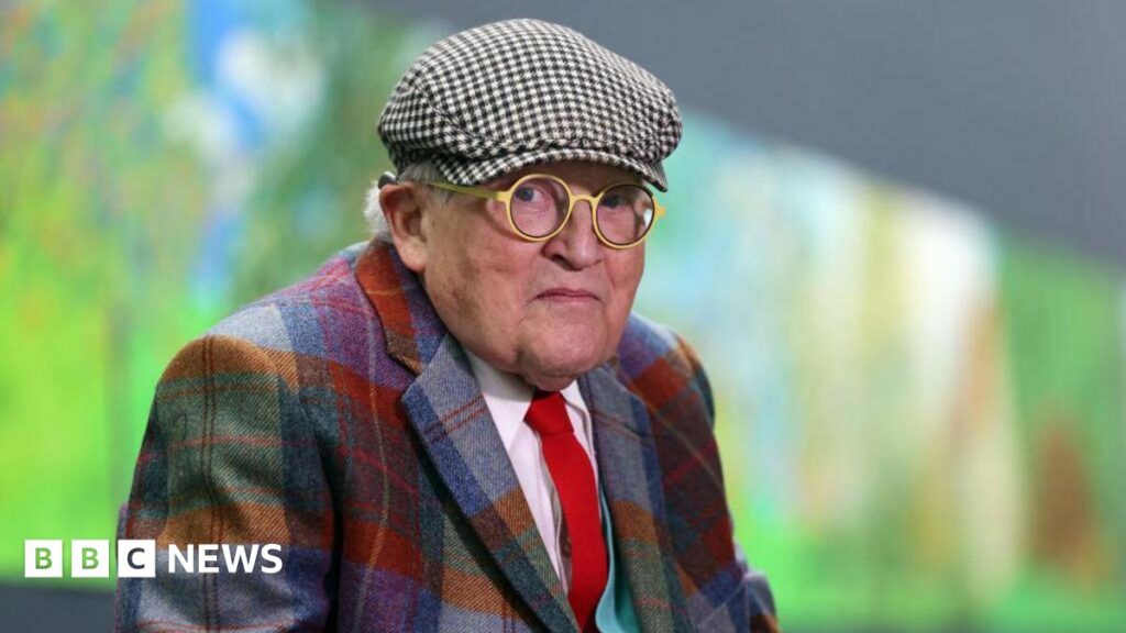 David Hockney says moving Bayeux Tapestry to UK is ‘madness’ David Hockney, wearing round glasses and a flat cap, along with a multicoloured checked shirt, in front of an artwork that can be seen blurred in the background
