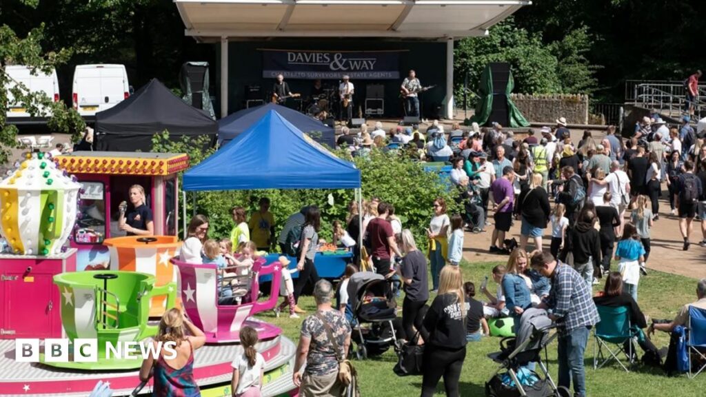 A small summer festival on a sunny day. Crowds of people including parents with prams sit or stand on a patch of grass in front of a stage where a band is performing. A spinning teacup ride is visible in the foreground.